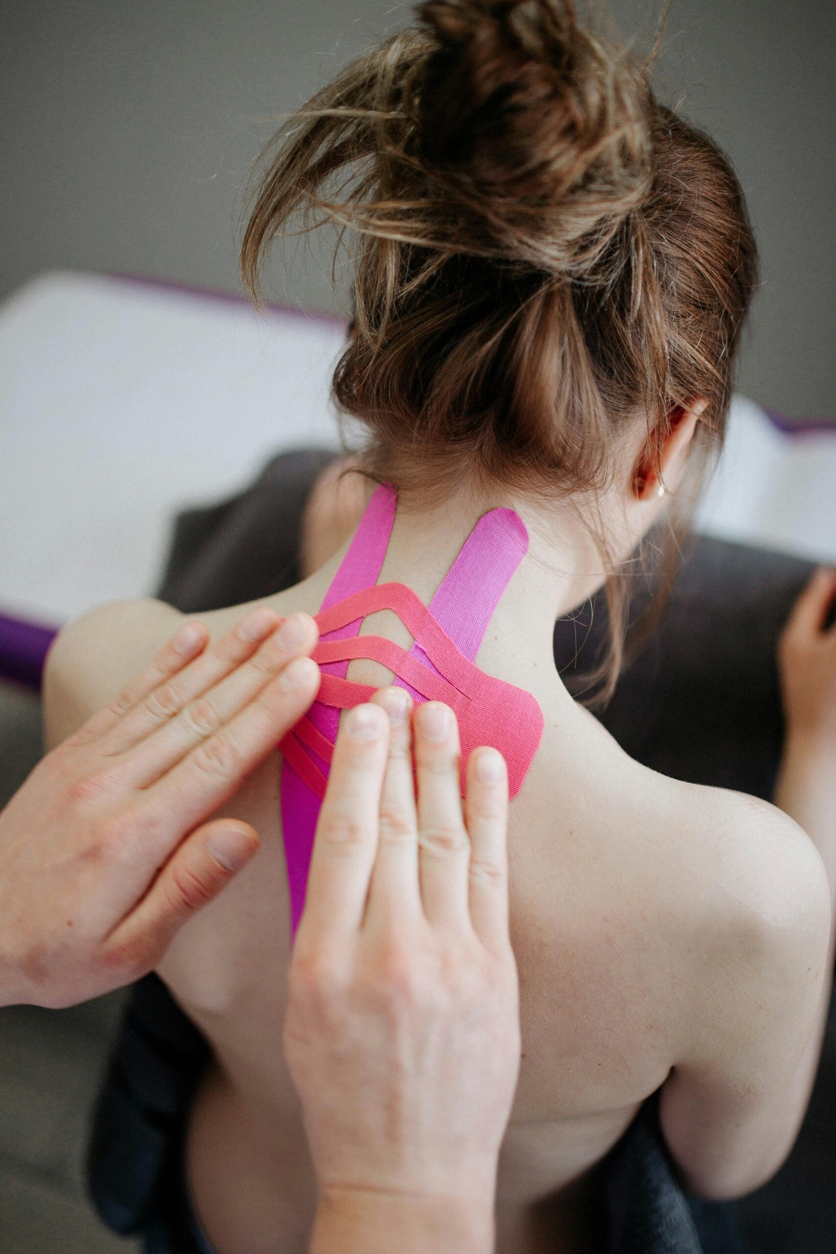 A pair of hands putting pink kinesiology taping onto the back of a woman's neck.